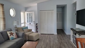 A living room with a grey sofa, a wooden coffee table, and a television on a cabinet. There is a door leading to a closet and a window with curtains. - Relaxing Getaway on the Island.  (Cape May)