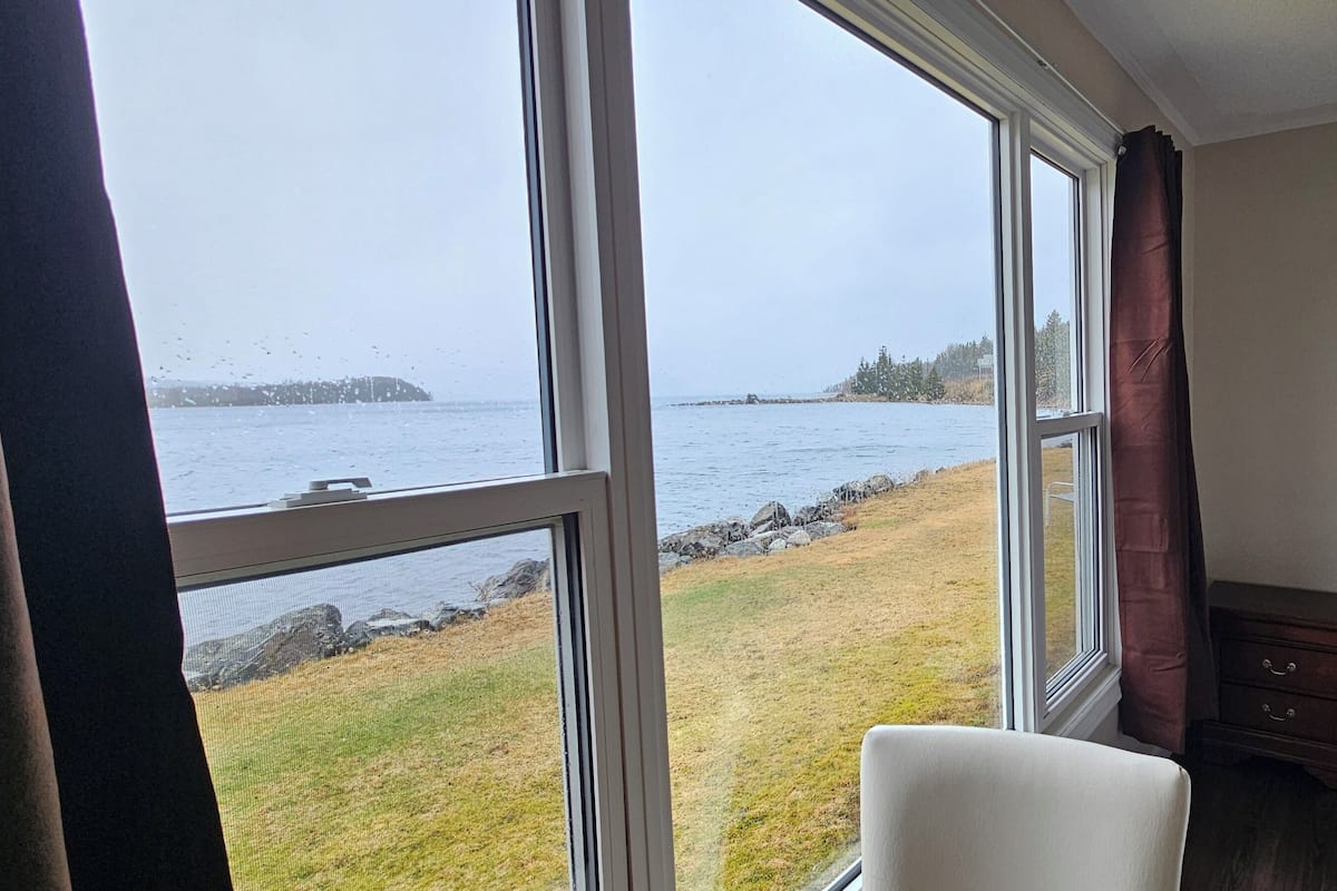 A bedroom with a large window overlooking a body of water, a bed, and a small bedside table.