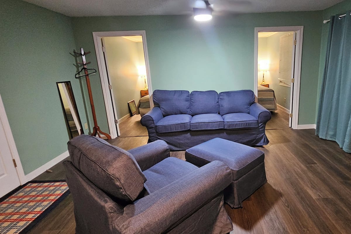 A living room with a sofa and armchair, a wooden floor, and a door leading to another room.