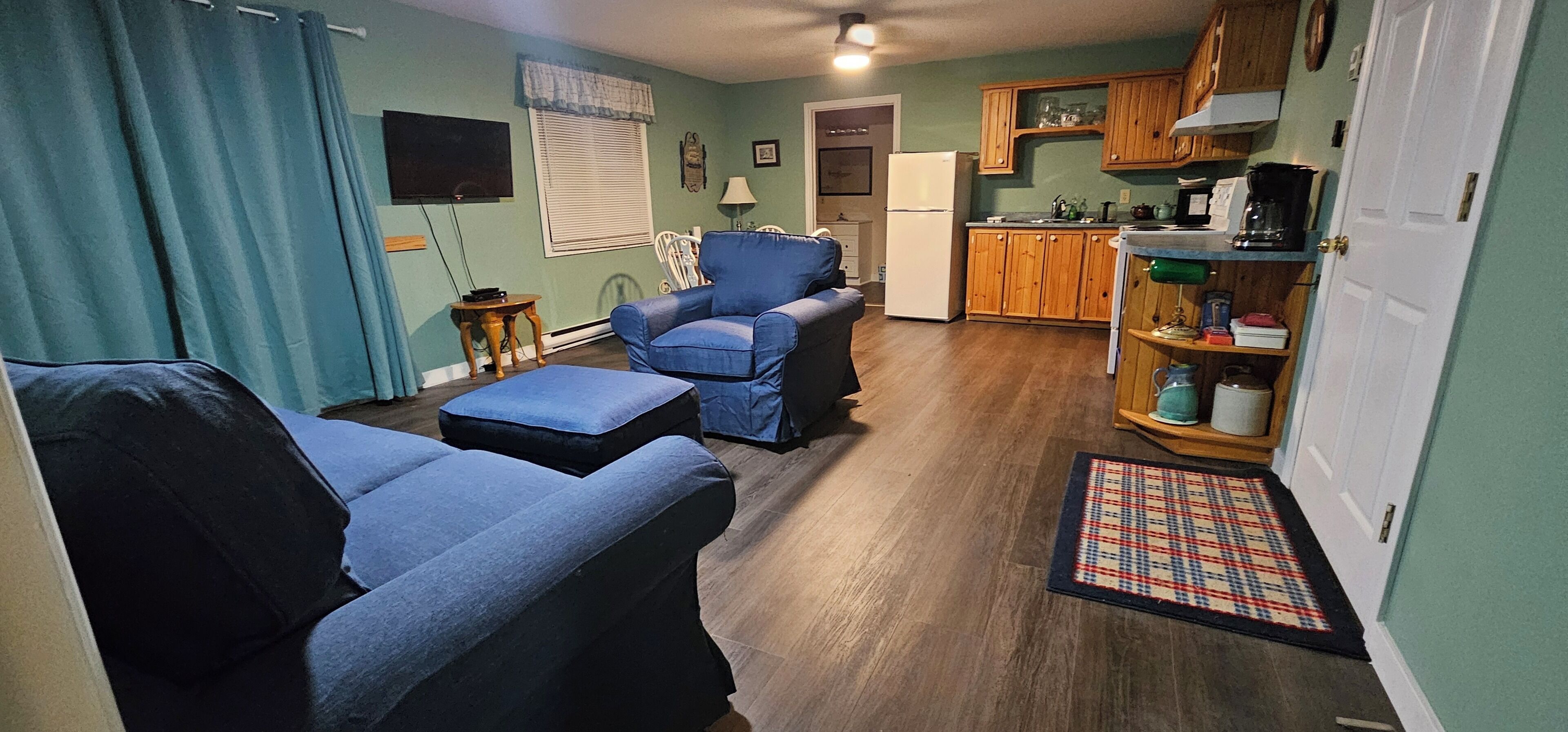 A compact living space with a sofa, a blue armchair, a small table, a kitchen area with wooden cabinets, and a television mounted on the wall.