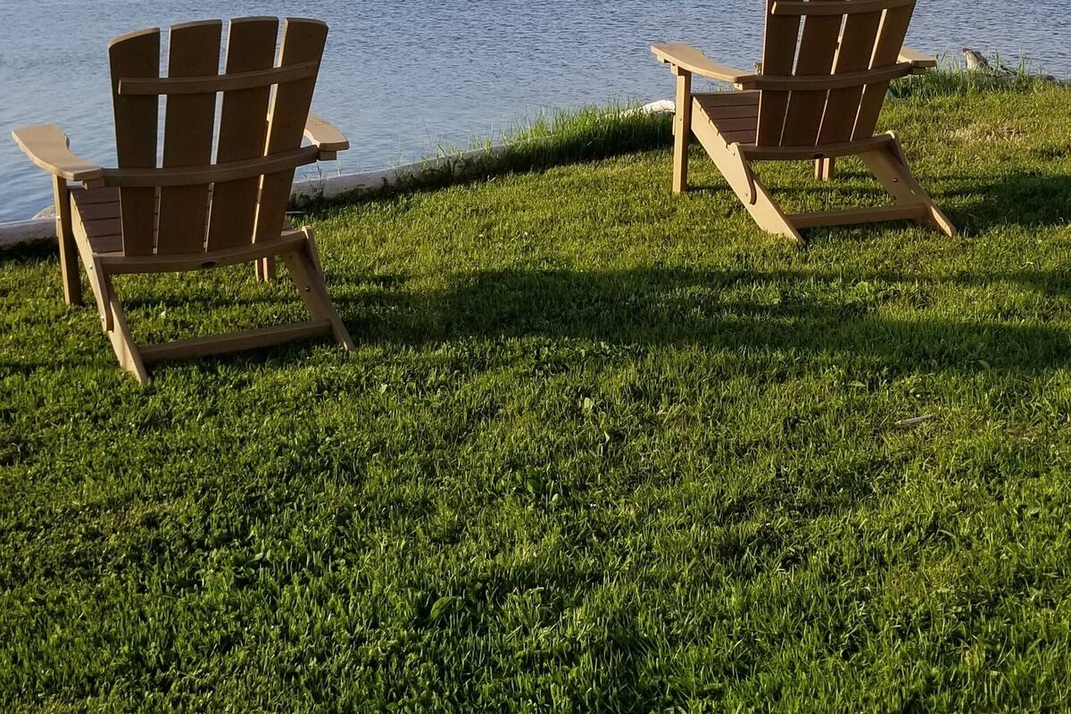 Two Adirondack chairs facing a lake with a grassy foreground and distant hills.