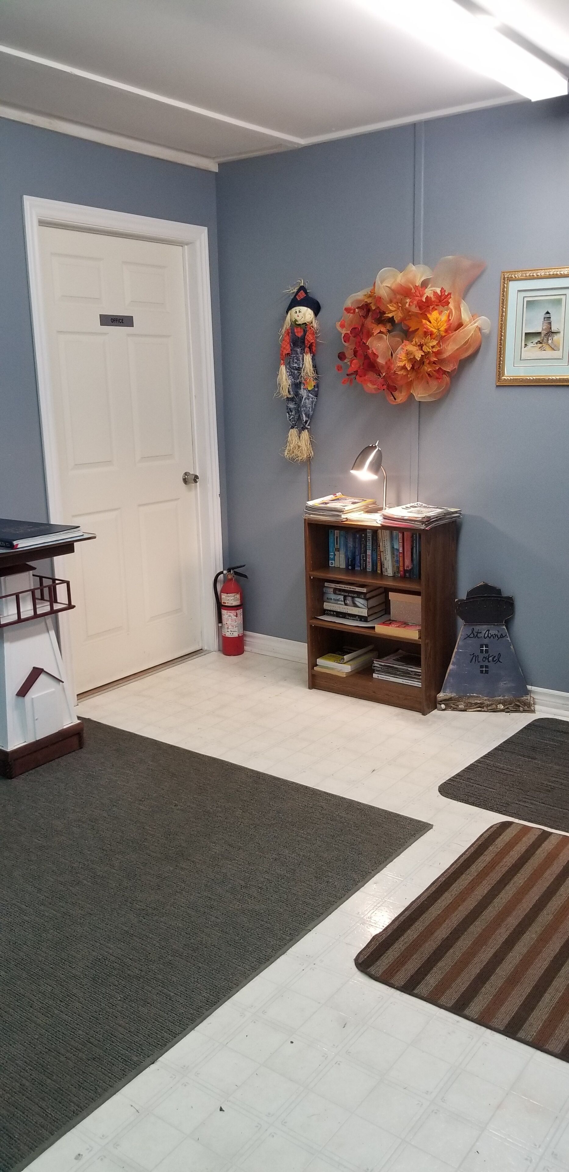 Una habitación con una puerta blanca que dice "OFFICE", una estantería con libros, un adorno en la pared y una mesita con una lámpara.