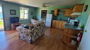 Sala de estar con sofá, televisor y mesa de comedor. Hay una cocina con armarios de madera y un refrigerador. Una ventana ofrece vistas al océano.