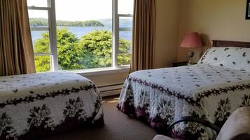 A hotel room with two beds, a window with a view of a lake and forest, a lamp, and a framed picture on the wall.