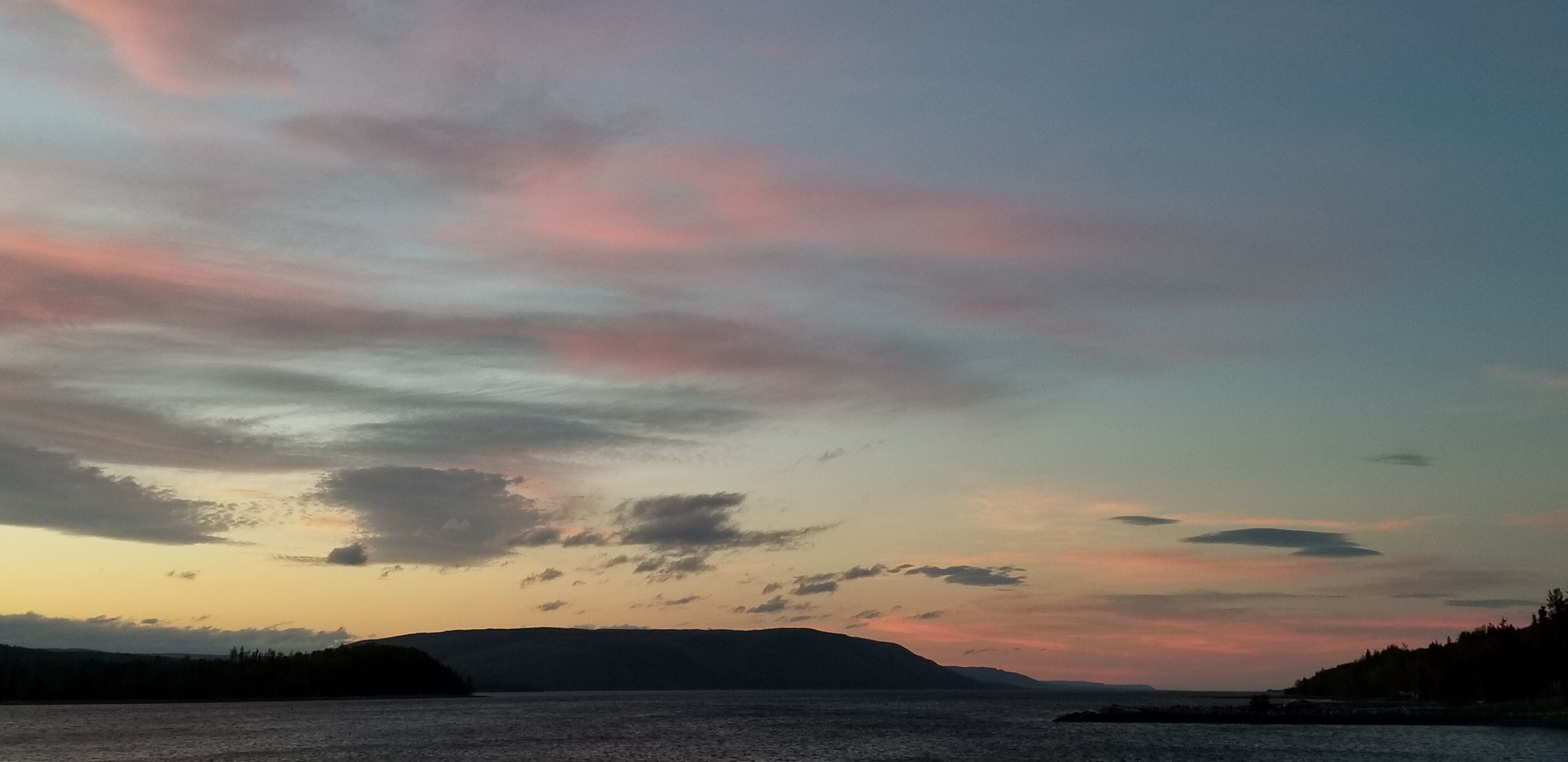 Un paysage côtier avec une étendue d’eau, des collines lointaines et un ciel au coucher du soleil.