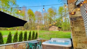 A hot tub on a stone patio with a wooden deck, surrounded by a stone wall and a grassy yard with trees. - Peaceful Retreat near Ohiopyle and Fallingwater HOT TUB (Mill Run)
