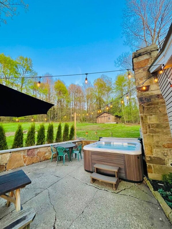 A hot tub on a stone patio with a wooden deck, surrounded by a stone wall and a grassy yard with trees.