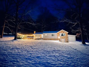 A snow-covered landscape with a house featuring a stone exterior, a garage, and a porch area with string lights.