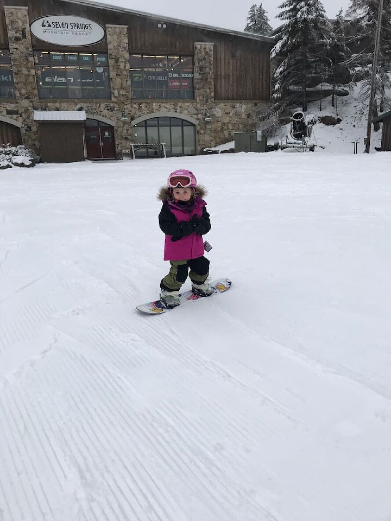 Un bambino fa snowboard davanti a un edificio di un complesso turistico in montagna.