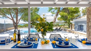 Une table à manger dressée pour six personnes, avec des serviettes, des assiettes et des couverts bleus, un vase rempli de fleurs et une vue sur une piscine et des arbres.