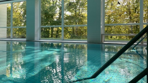 Ein heller, überdachter Pool mit klarem, blauem Wasser, großen Fenstern und Blick auf Bäume draußen.