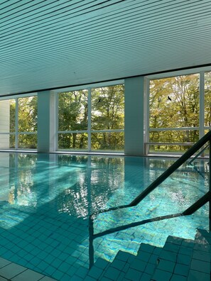 Ein heller, überdachter Pool mit klarem, blauem Wasser, großen Fenstern und Blick auf Bäume draußen.