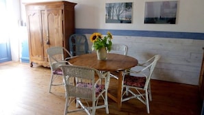 A dining area with a round wooden table surrounded by wicker chairs, a wooden cabinet, and two framed pictures on the wall. - Typical house in Saint Sauveur (L'Île-d'Yeu)