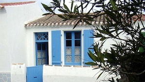 A white building with blue shutters and a blue door, featuring a tiled roof and a small porch area. - Typical house in Saint Sauveur (L'Île-d'Yeu)