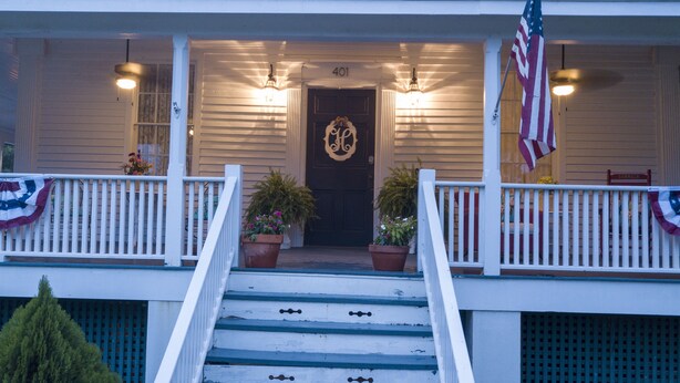 Uma casa de dois andares com varanda frontal, uma porta preta com emblema decorativo e uma bandeira americana.