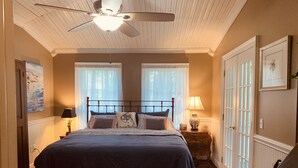 Um quarto com uma cama, duas janelas com cortinas leves, um ventilador de teto, uma mesa de cabeceira com abajur e uma porta com painéis de vidro.