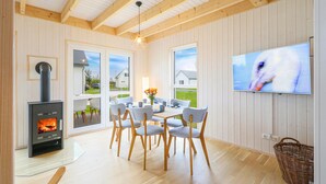 Una sala moderna con estufa de leña, comedor con sillas y mesa, televisor montado en la pared y ventanas amplias con vistas al exterior.