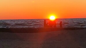 Een zonsondergang boven de zee, met silhouetten van mensen op het strand.