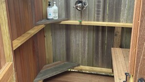 A wooden outdoor shower with a showerhead and hose, a small shelf with toiletries, and a bench.