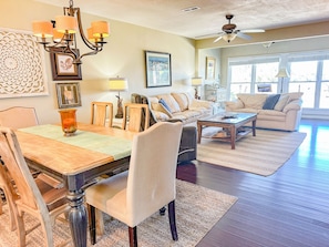 A spacious living room with a dining area, featuring a chandelier, a ceiling fan, and large windows.