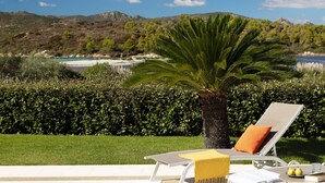 A poolside lounge chair with a yellow towel and an orange cushion, a palm tree, and a view of a beach and mountains in the background. - VILLA IPPOCAMPO private pool and close to the sandy beach (Capo Coda Cavallo)