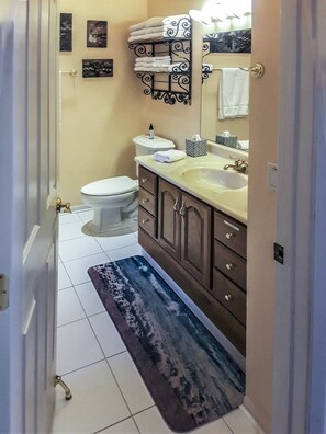 A bathroom with a toilet, sink, and vanity. There are towels stacked on a shelf, a rug on the floor, and a mirror above the sink.