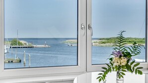 Uitzicht vanuit een raam op een jachthaven met aangemeerde boten, een heldere lucht en een plant in een vaas op de vensterbank.