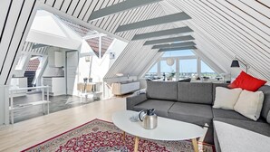 Een modern loft appartement met een schuine plafond, een gezellige zithoek met een grijze bank en een rood kussen, een witte salontafel en een vloerkleed met patroon.