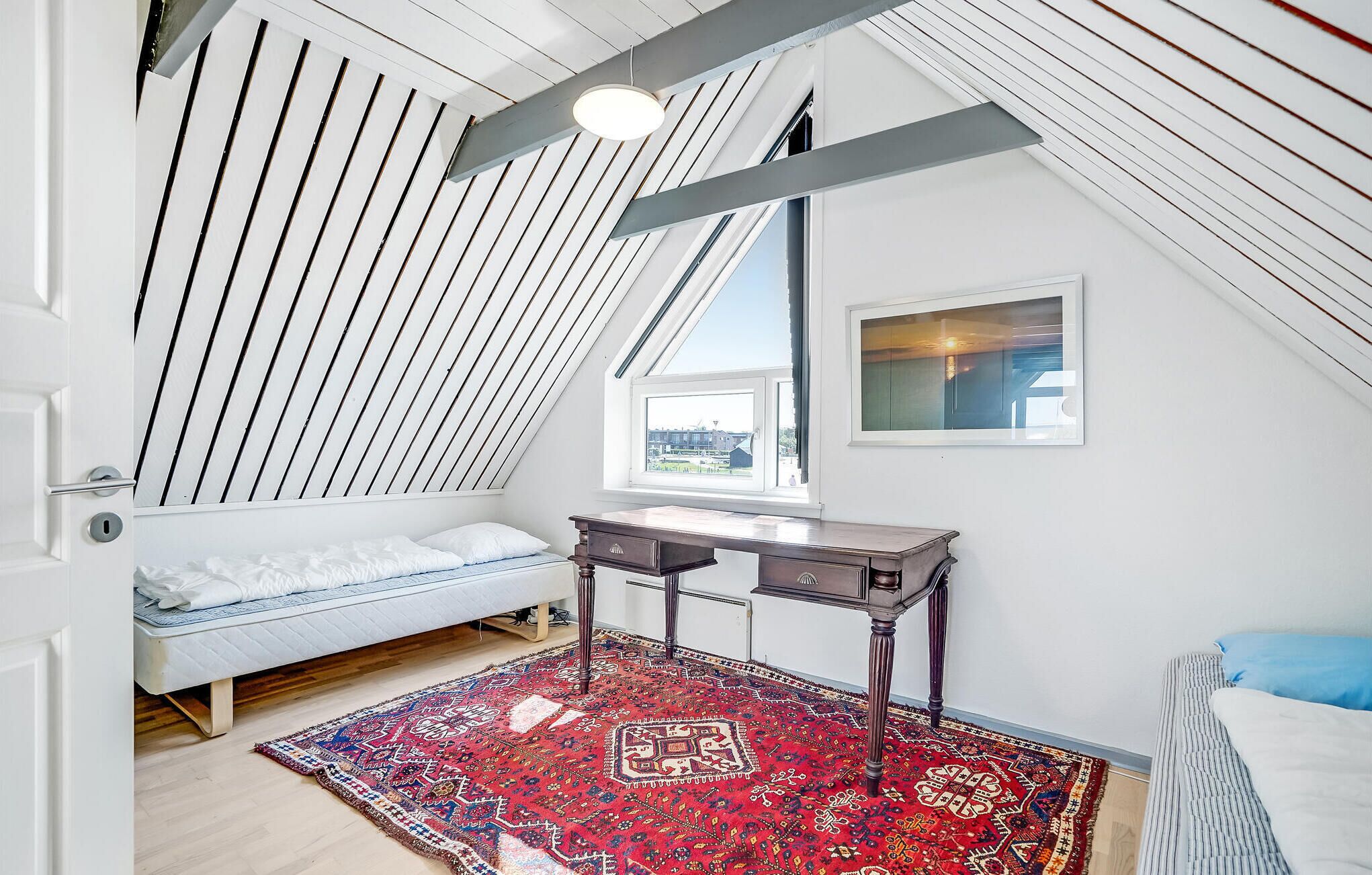 A bedroom with a sloped ceiling, a bed, a desk, a window, and a view of the outside.