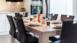 A modern dining area with a wooden table set for six, black chairs, and a kitchen in the background. - Villa Dalu am See - 5* luxury vacation home (Göhren-Lebbin (OT Untergöhren))