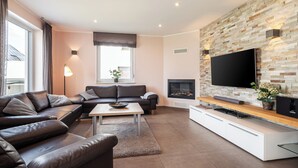 A modern living room with a sectional sofa, a coffee table, a flat-screen TV mounted on a stone wall, and a fireplace. - Villa Dalu am See - 5* luxury vacation home (Göhren-Lebbin (OT Untergöhren))