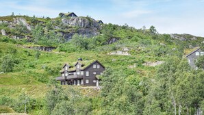 Una casa grande de madera oscura con varios pisos y un balcón destacado, ubicada en una ladera cubierta de hierba, rocas y abundante vegetación.