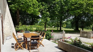 An outdoor patio with a wooden table and chairs, surrounded by a garden with flowers and a fence. - Pretty stone house with swimming pool in magnificent agricultural area (Bonnes)