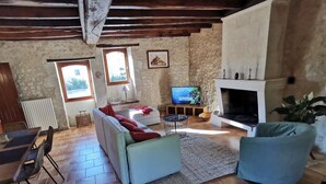 A rustic living room with exposed wooden beams, a stone wall, a fireplace, a television, and a sofa with red cushions. - Pretty stone house with swimming pool in magnificent agricultural area (Bonnes)