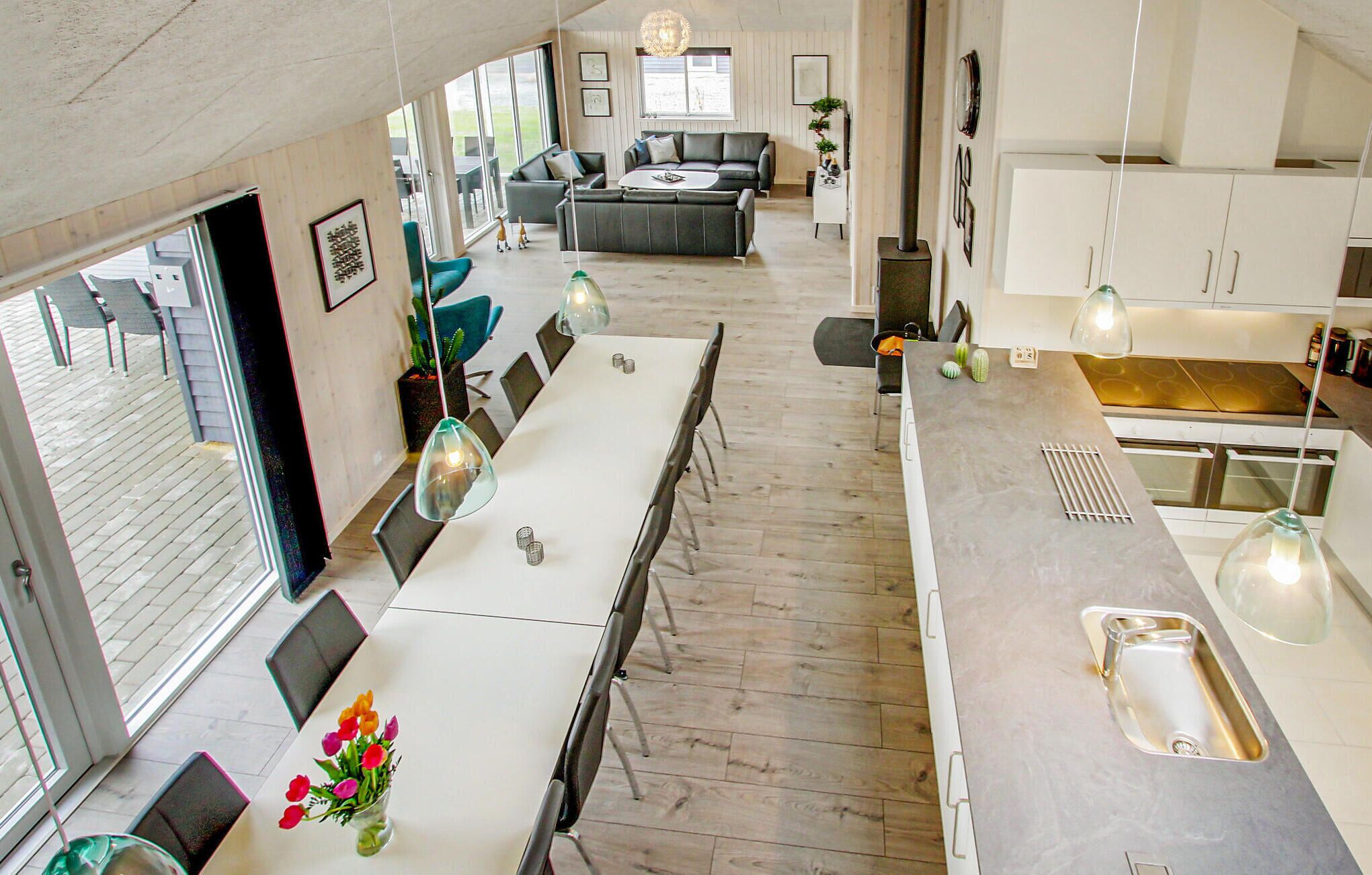 Een moderne keuken met een lange aanrecht, een spoelbak en hanglampen. Er is een eethoek met een lange tafel en stoelen, en een zithoek met een bank en salontafel.