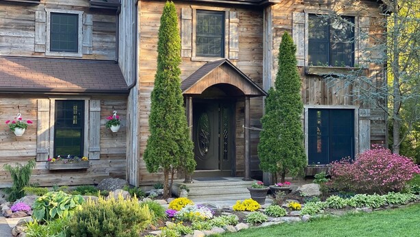 A two-story wooden house with a front porch, a small garden with various plants and flowers, and a large window on the second floor. - Unique home in great neighborhood (Stroudsburg)