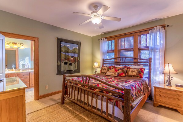 A bedroom with a wooden bed, a ceiling fan, a window with blinds, a nightstand, and a mirror.