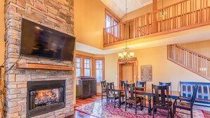 A spacious living room with a stone fireplace, a dining area with wooden furniture, and a balcony with wooden railings. - Bearfoot Lodge at Echota Resort - Pool, Gym, Fishing Access, Private hot tub (Boone)