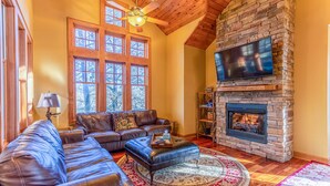 A cozy living room with a stone fireplace, leather sofas, and a large window. - Bearfoot Lodge at Echota Resort - Pool, Gym, Fishing Access, Private hot tub (Boone)