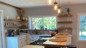 Cocina moderna con una isla central, taburetes de bar, iluminación colgante y vistas a la vegetación.
