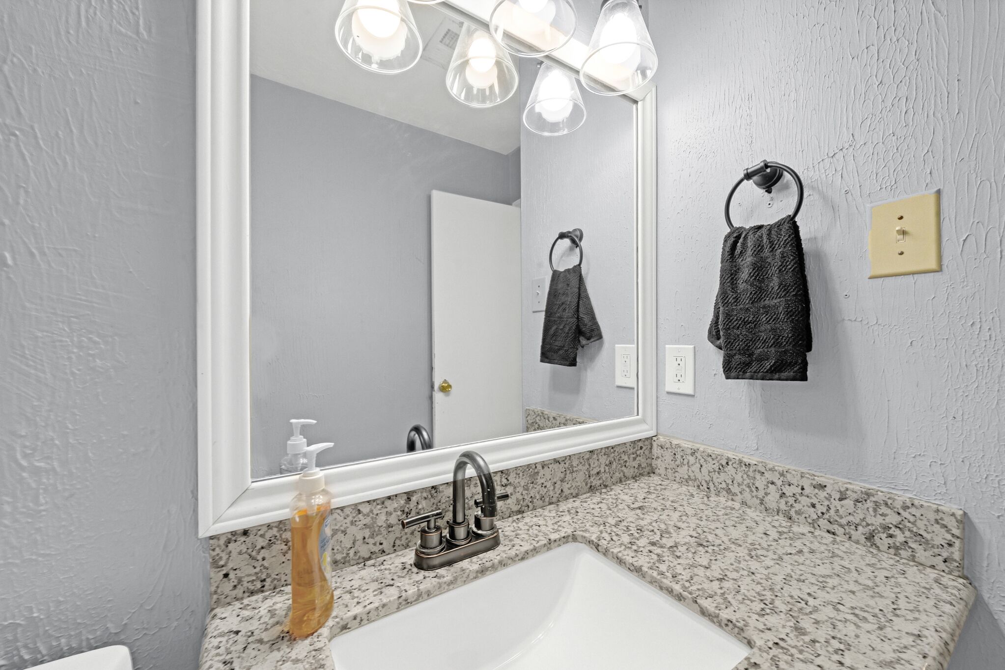 Een badkamer met een granieten aanrecht, een witte wastafel, een grote spiegel en een handdoekhouder met twee handdoeken.