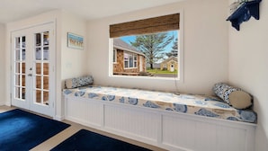 Quarto com um banco embutido na parede, janela com vista para uma casa e porta com painéis de vidro.