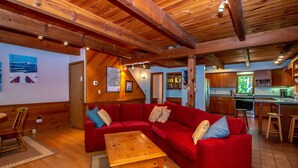 A cozy living room with a red sofa, wooden coffee table, and a kitchen area with wooden cabinets and a bar stool. - Trailside off West Side! (North Conway)