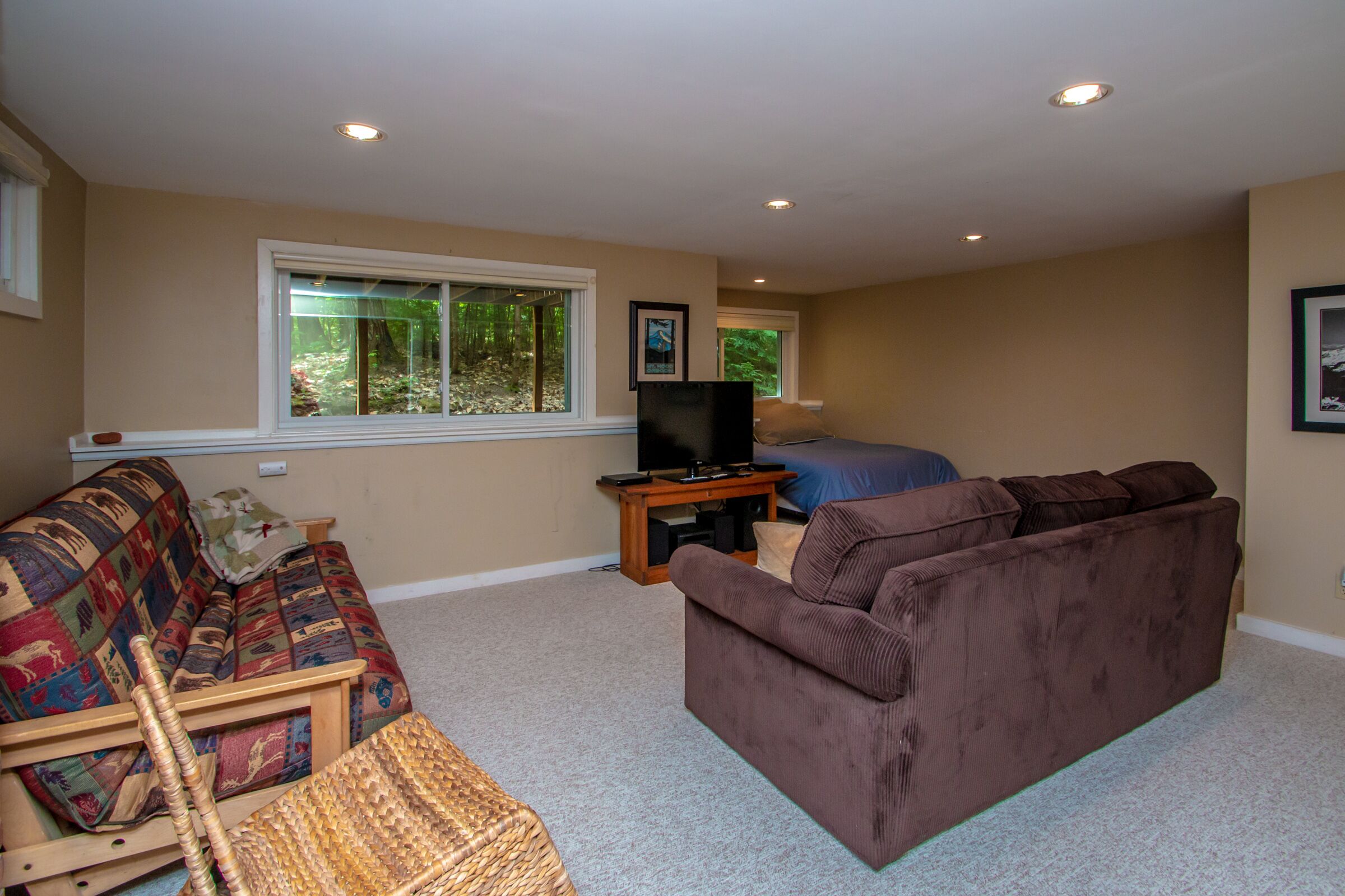 A living room with a brown sofa, a colorful quilted couch, a wooden chair, a television on a stand, and a window with a view of trees.