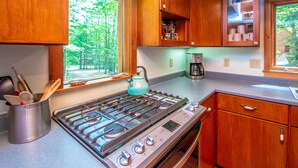 A kitchen with wooden cabinets, a gas stove, and a window overlooking a forest. - Trailside off West Side! (North Conway)