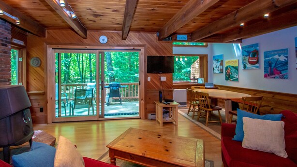 A cozy cabin interior with a wooden ceiling, a fireplace, a dining area, and a large sliding glass door leading to an outdoor deck. - Trailside off West Side! (North Conway)