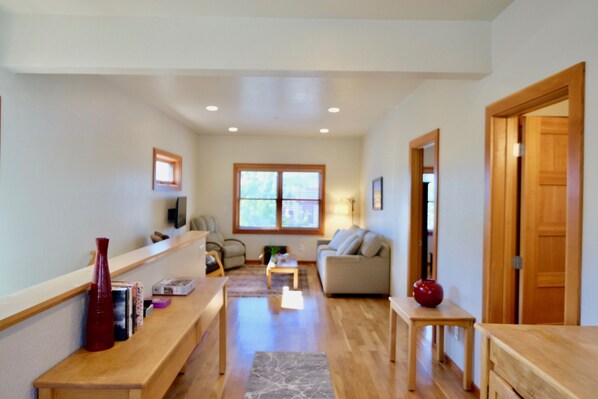 A spacious living area with a sofa, a coffee table, and a wooden bench. There is a window with blinds, a door, and a small table with a vase.