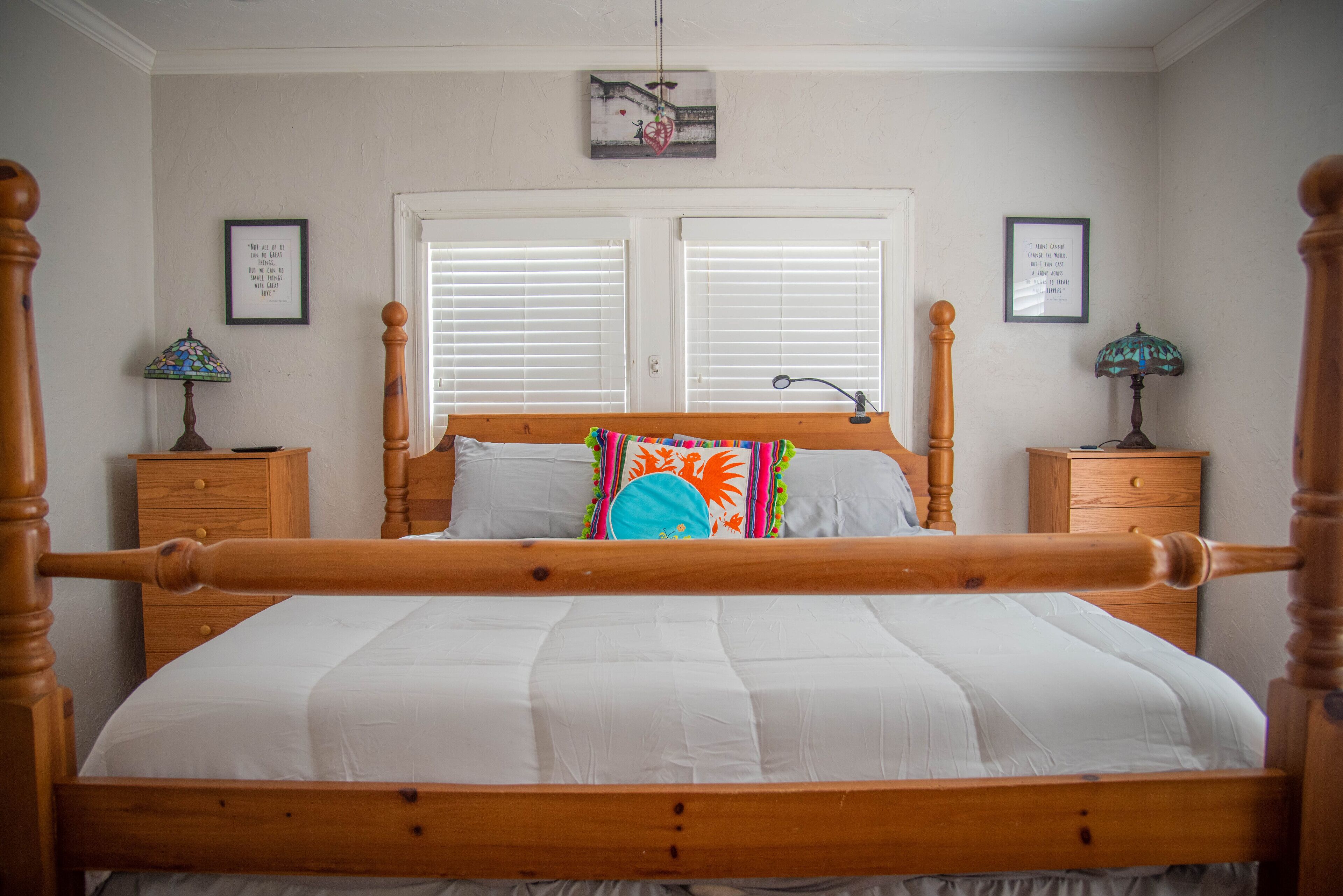 A bedroom with a wooden four-poster bed, white bedding, two nightstands with lamps, and a window with blinds.