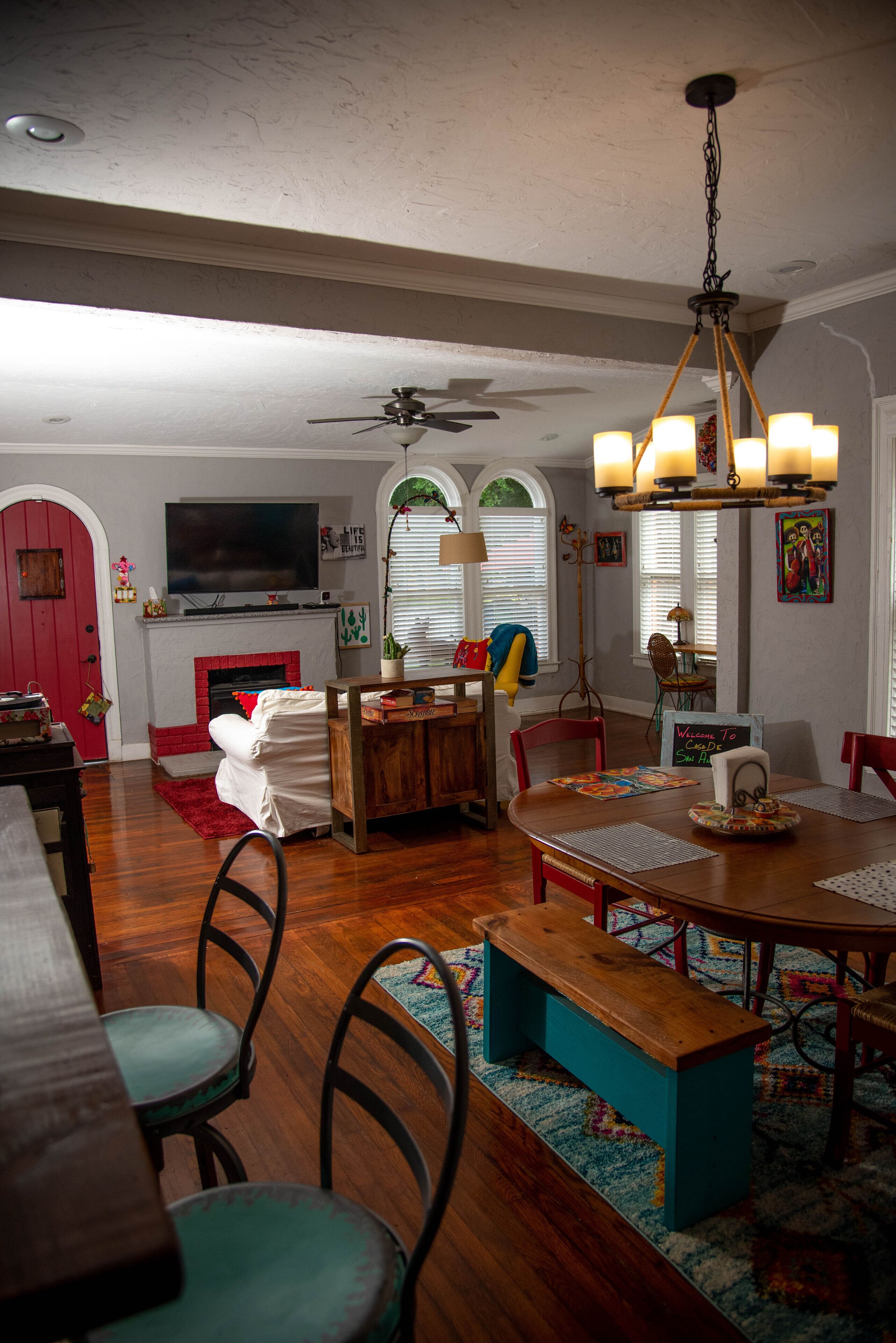 A dining area with a wooden table, chairs, and a bench. There is a fireplace with a red door, a television mounted above it, and a ceiling fan.