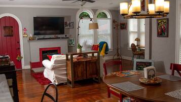 A dining area with a wooden table, chairs, and a bench. There is a fireplace with a red door, a television mounted above it, and a ceiling fan.